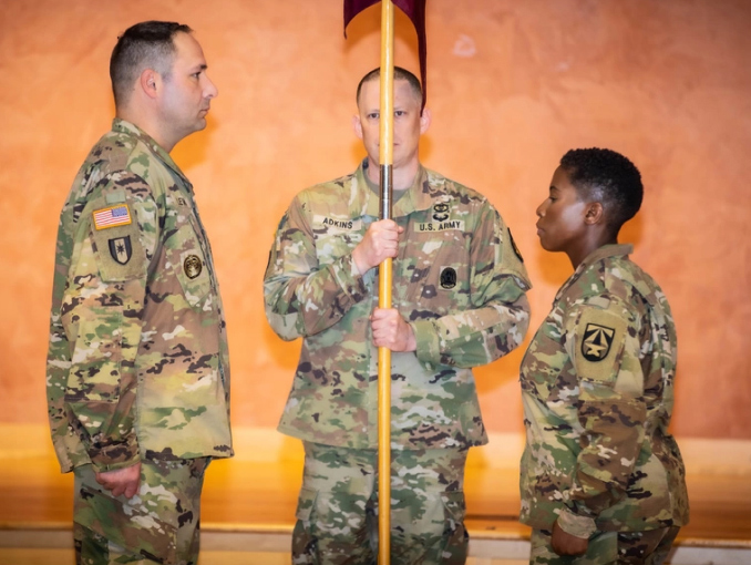 Una Soldado y dos Soldados en uniforme de combate están en posición de pasar una bandera durante una ceremonia