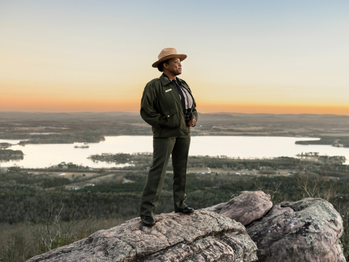 A female park ranger standing on top of a mountain overlooking a lake at sunset