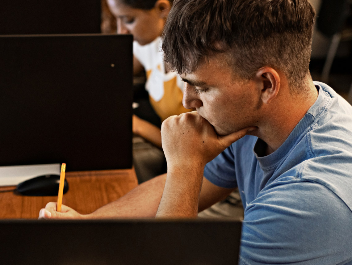 A male enlisted recruit wearing a light blue t-shirt sitting down at a brown desk and writing with a pencil