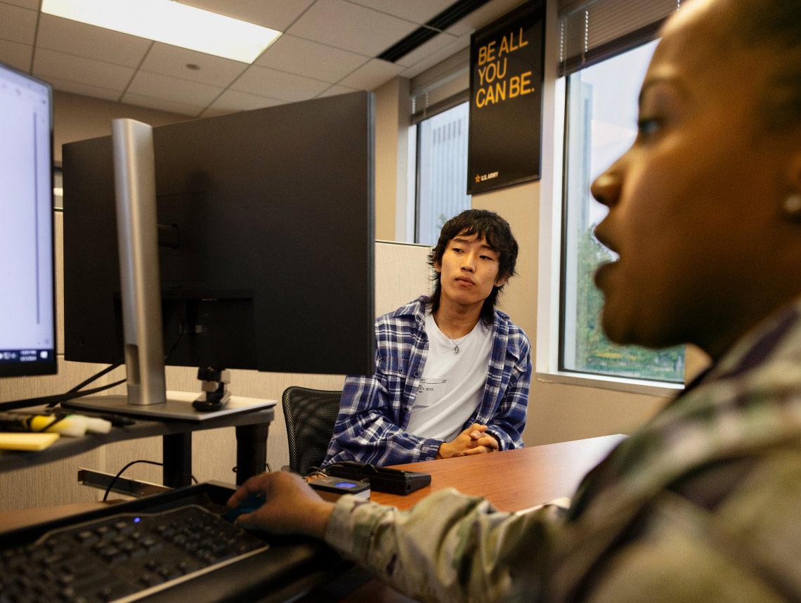 An enlisted recruit and Soldier sitting down at a brown desk and staring at a computer on the desk