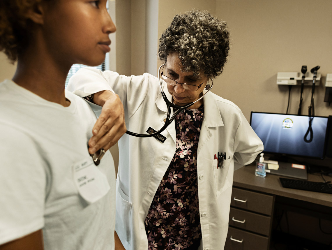 A female doctor in a white uniform placing a stethoscope on the chest of a female enlisted recruit