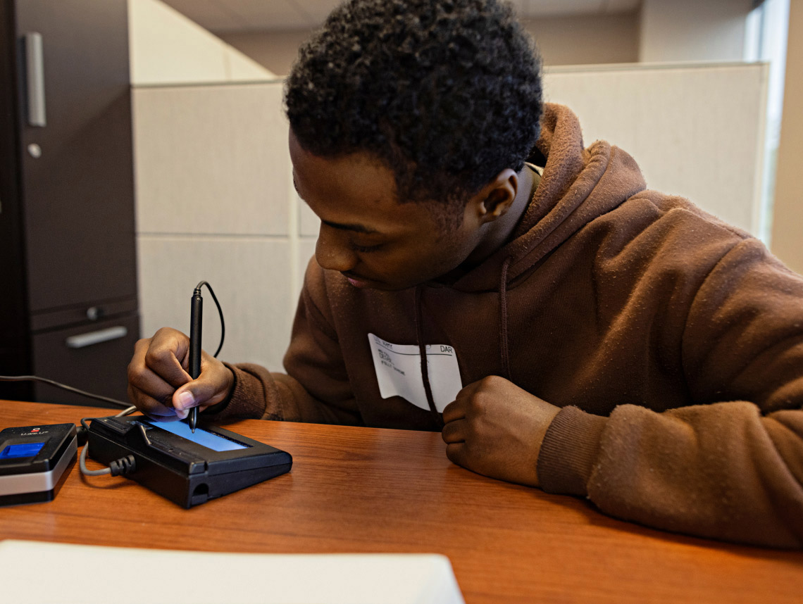 A male enlisted recruit wearing a brown sweatshirt, sitting down at a brown desk and writing his electronic signature