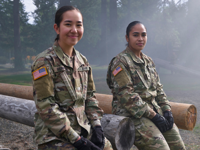 Two female ROTC cadets sitting outside against a wooden obstacle