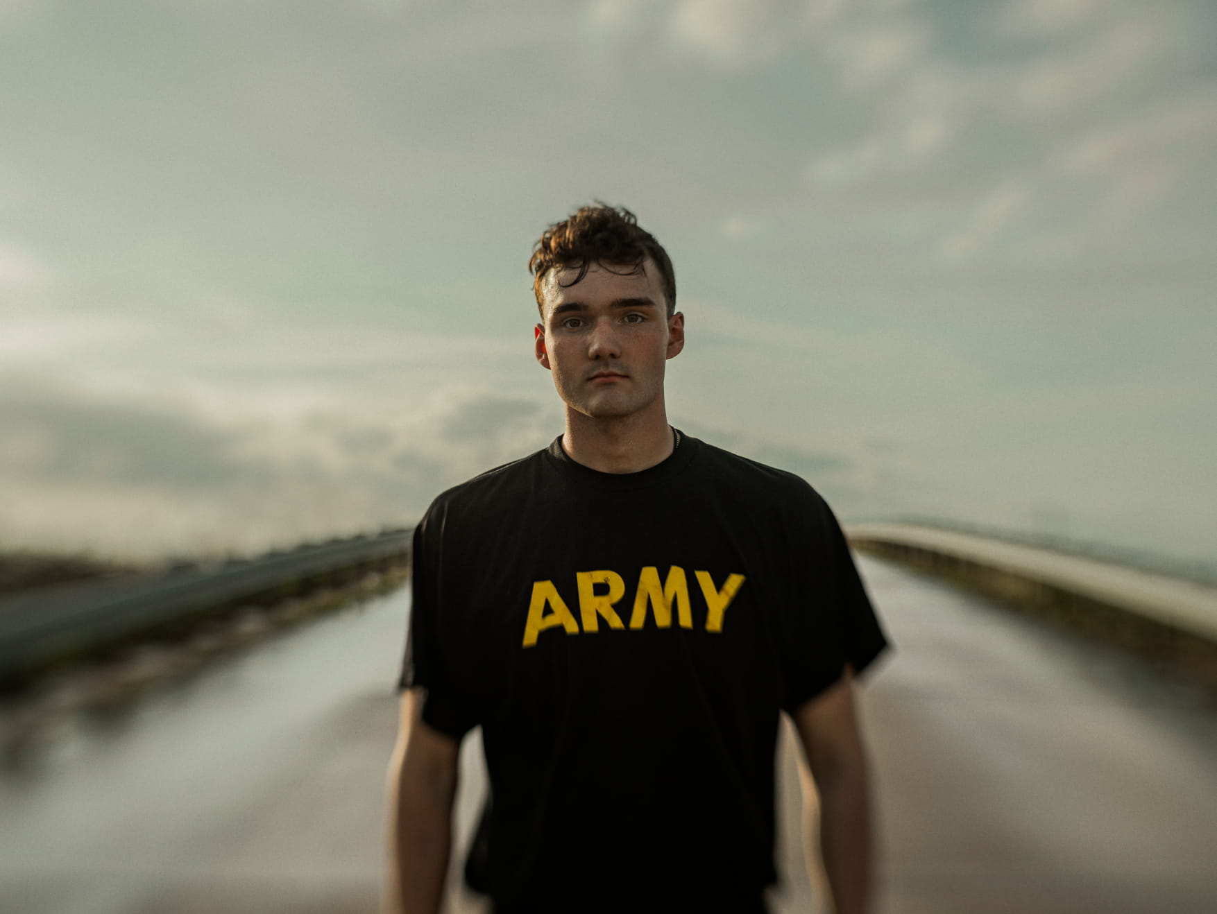 Young man wearing a black Army T-shirt stands on a bridge at sunset