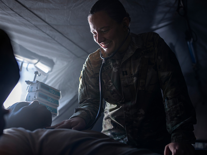 Female Army Medicine doctor smiling while holding a stethoscope onto the chest of a limp patient