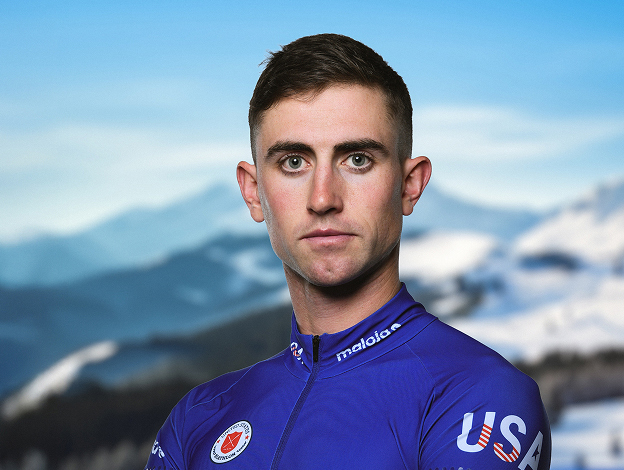Specialist Sean Doherty wearing a blue USA uniform and looking at camera with arms crossed and snowy mountains in the background