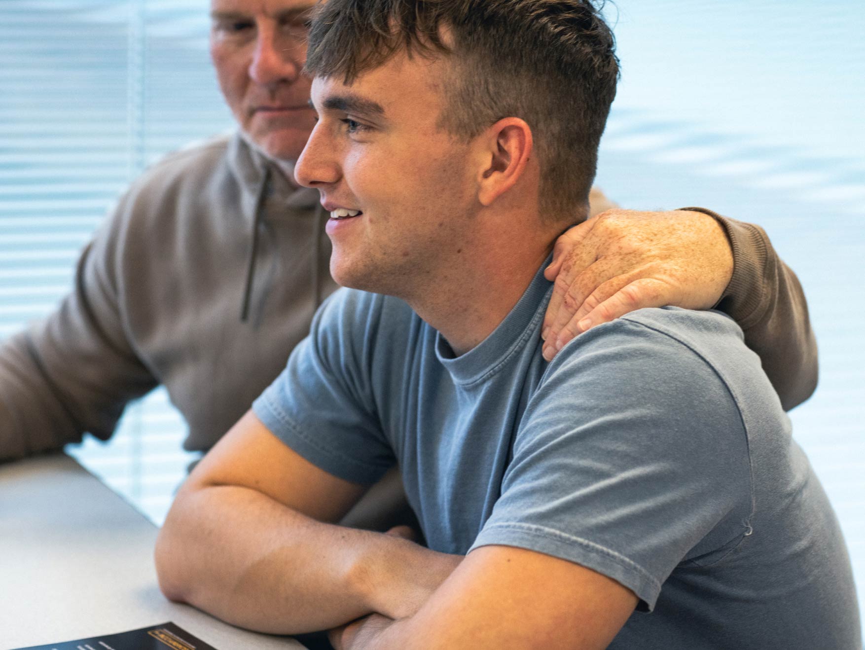 A male enlisted recruit wearing a light blue t-shirt, sitting down and staring into the distance with another person’s arm wrapped around his shoulder