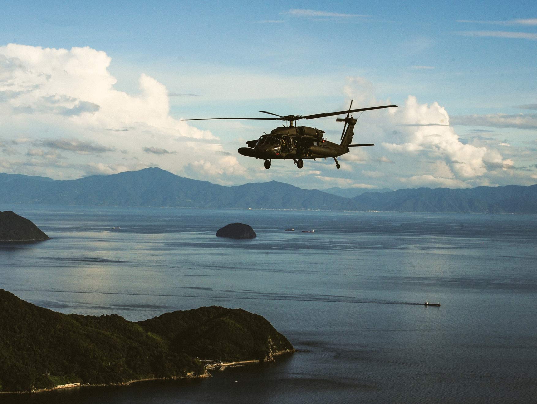 An Army Black Hawk helicopter flying over a large body of water with mountains and bright cloudy skies in the background.