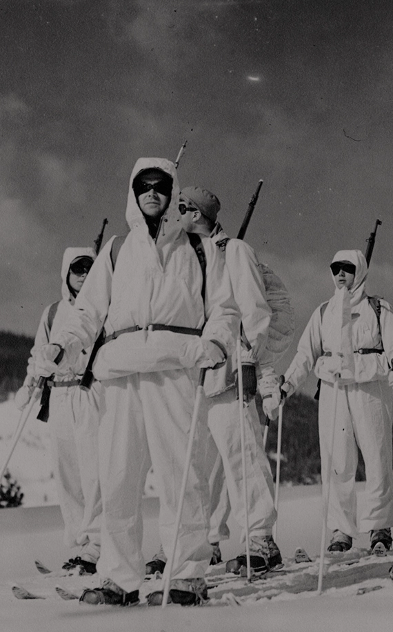 Hombres de la 10th Mountain Division de pie sobre esquís y vistiendo uniformes blancos con gafas de sol, mientras miran fijamente a la distancia