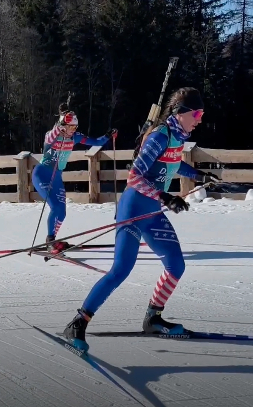 Staff Sergeant Deedra Irwin usando un uniforme rojo, blanco y azul de biatlón mientras realiza esquí de fondo con bastones de esquí en la mano y un rifle en la espalda