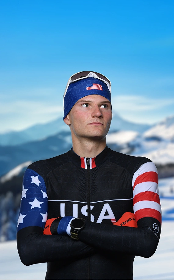 Sergeant Benjamin Loomis in a black, red, white, and blue USA ski uniform, crossing arms looking to the side with snowy mountains in the background