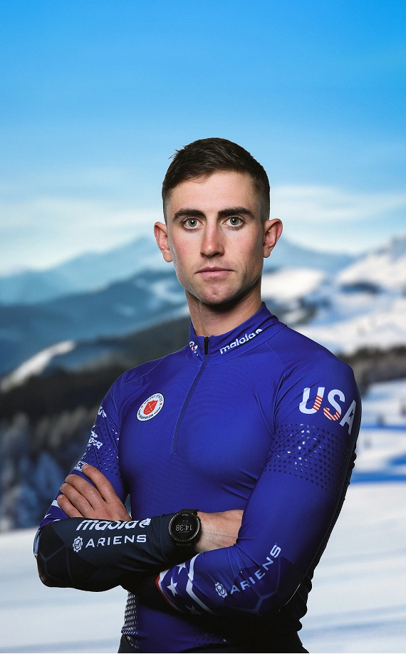 Specialist Sean Doherty wearing a blue USA uniform and looking at camera with arms crossed and snowy mountains in the background