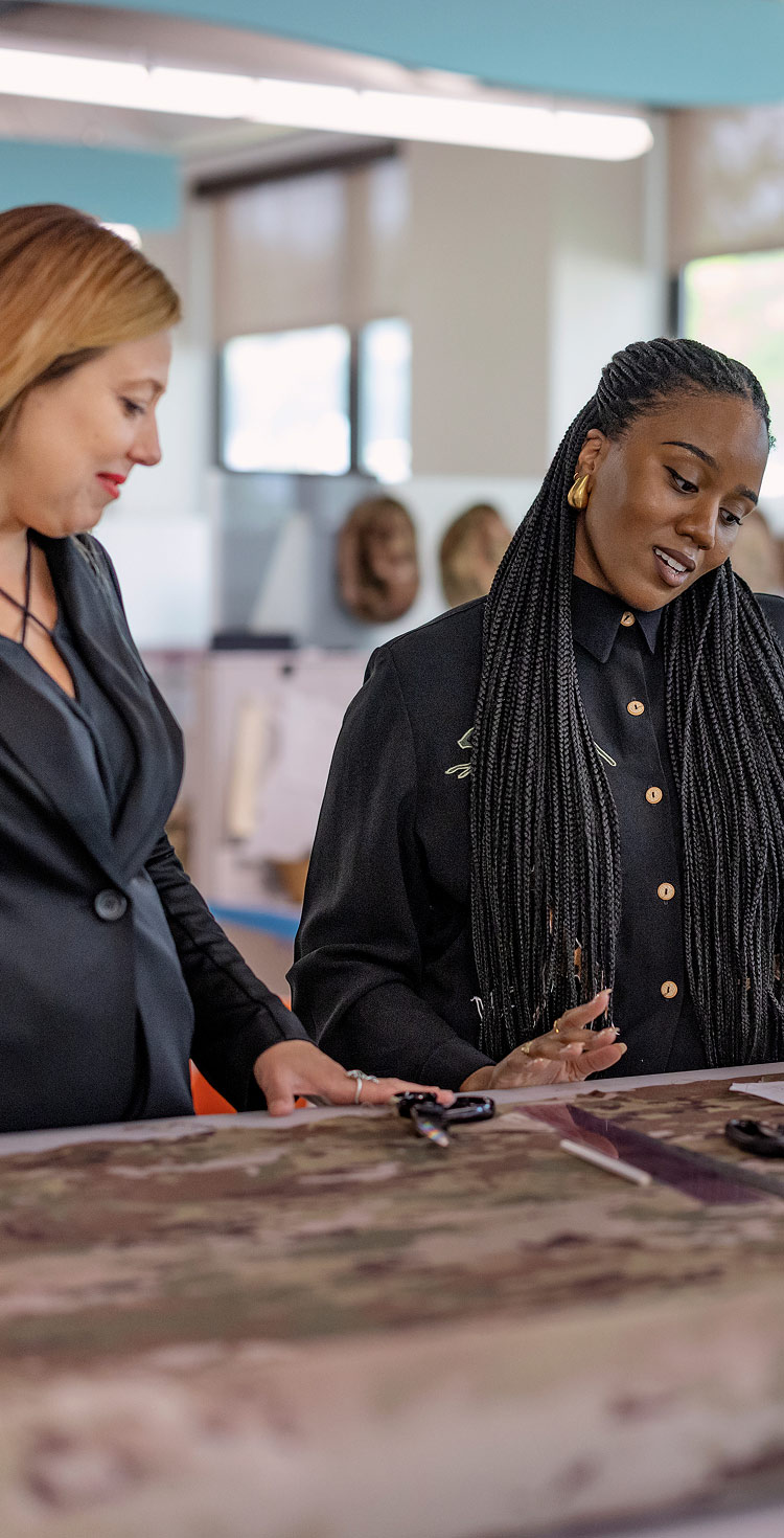 Two clothing designers talking while standing in front of a work desk with scissors and camouflage fabric 