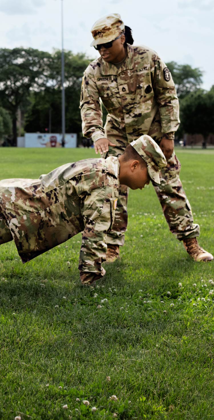 A drill sergeant standing over a group of enlisted recruits on a grass surface, who are all focused on completing a push-up