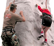 A cadet coaches a young girl up a rock climbing wall.